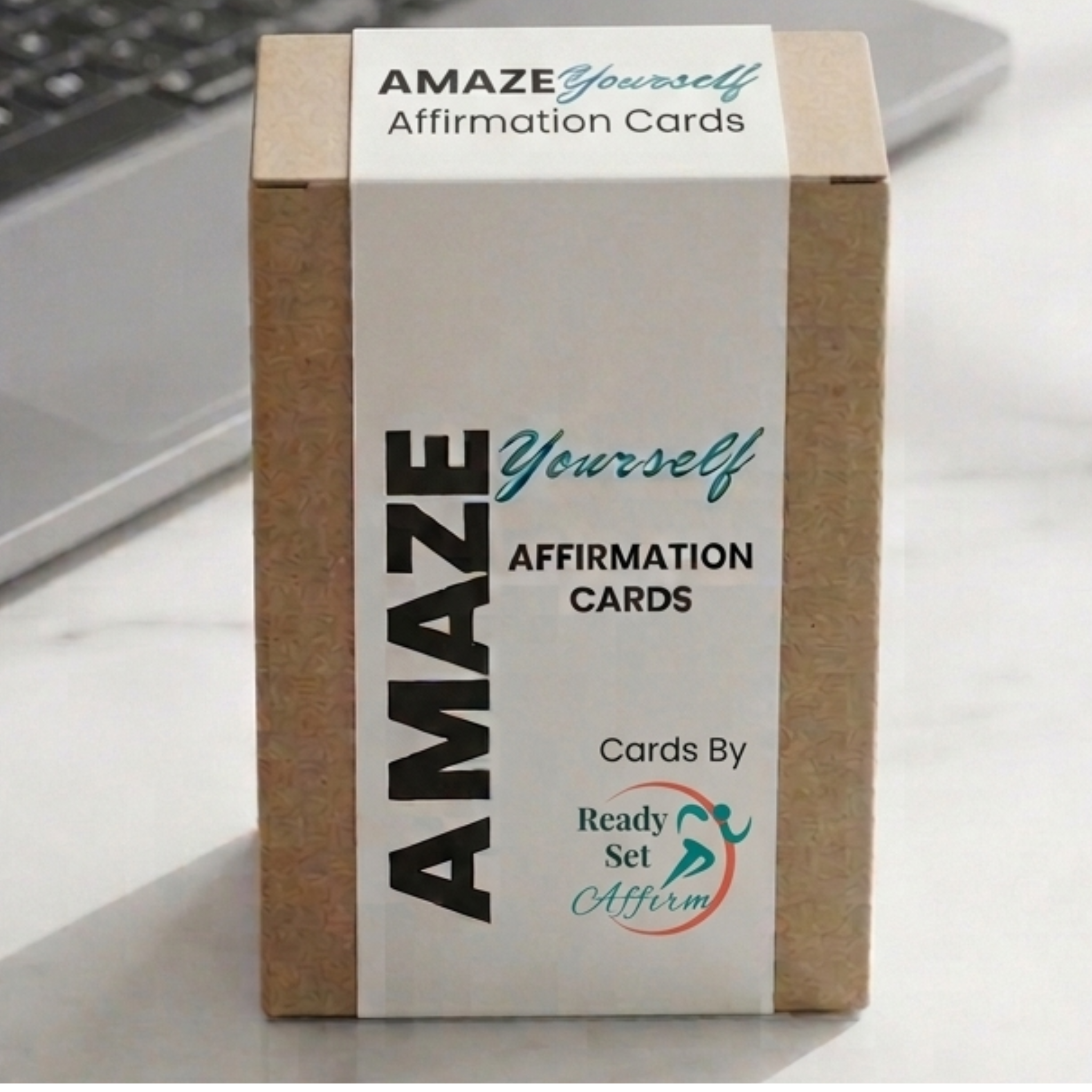 A professional workspace desk setup featuring the 'AMAZE Yourself Affirmation Cards' by Ready Set Affirm. The premium brown kraft paper product box is wrapped in a sleek, white vertical band displaying the teal and black brand logo. Positioned upright on a polished white marble office desk, a laptop rests softly out of focus, illuminated by warm, natural light to create a calm, high-performing workspace aesthetic