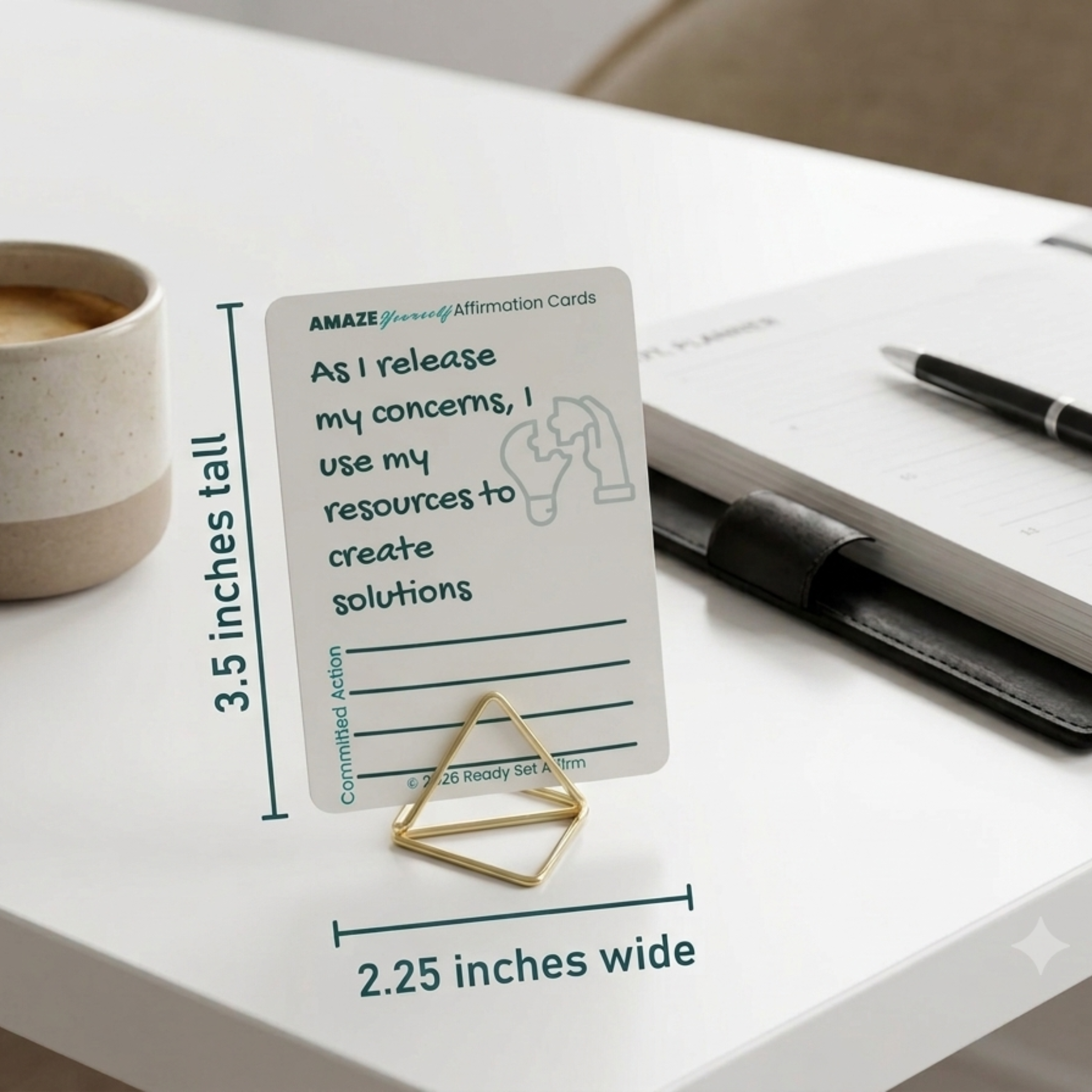 professional office desk setup featuring a single 'AMAZE Yourself' affirmation card resting in a modern, geometric gold wire card holder. The textured card displays the stress-management affirmation, 'As I release my concerns, I use my resources to create solutions' (card by Ready Set Affirm), with blank lines below for writing a committed Action for the day. Placed on a clean, bright white desk, the card stands next to a rustic ceramic coffee mug filled with fresh espresso. A sleek black pen and minimalist