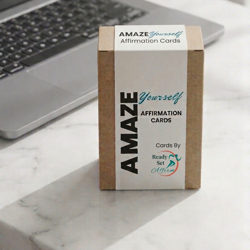 A box of AMAZE Yourself Affirmation Cards standing on a white marble desk next to a blurred laptop.