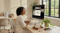 A woman sits at a desk in a bright home office, watching a virtual workshop on a computer monitor attending Ready Set Affirm's workshop, "The Restoration Protocol". The room has natural light from a large window, with plants, books, and a phone on the desk.